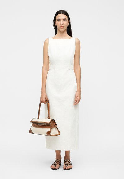 Γυναίκα με μακριά σκούρα μαλλιά φοράει λευκό μίντι φόρεμα χωρίς μανίκια, μαύρα σανδάλια με λουράκια και κρατάει μια λευκή και καφέ τσάντα με δύο λαβές.