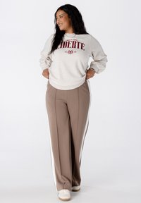 Femme debout souriante, portant un sweat-beige avec le texte "LIBERTE", un pantalon taupe avec des rayures blanches sur les côtés, et des baskets blanches sur fond blanc.