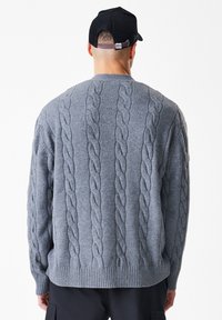 Siv knitani pulover z okroglim izrezom, teksturiranimi vzorci in rebrastim robom. Zadnji pogled prikazuje ohlapen kroj in debel pleten material.