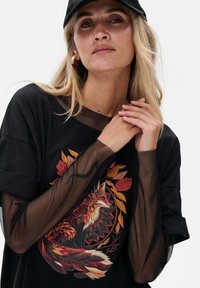 Zwarte t-shirt met een vosgrafiek, voorzien van rode, oranje en gele borduursels. Doorzichtige zwarte mazen mouwen voegen textuur en contrast toe.