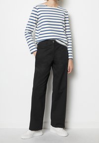 Gestreiftes Langarmoberteil in Weiß und Marineblau, kombiniert mit schwarzen weit geschnittenen Hosen. Das Model trägt weiße Sneakers. Minimalistisches Design und Passform.