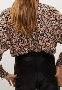 Blouse noire à motifs floraux avec des manches bouffantes présentant des fleurs orange et blanches ; portée avec un pantalon en cuir noir taille haute. Texture lisse.