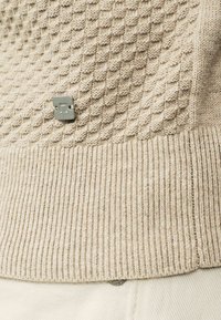 Beige sävyinen neulepaita, jossa on mehiläiskennokuvio ja resorireuna, sekä pieni metallinen logo rintakehässä.