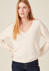 Cremfarbener, strukturierter Strickpullover mit weiter V-Ausschnitt, langen Ärmeln und entspanntem Schnitt, kombiniert mit dunklen Jeans. Dezente vertikale Rippendetails.