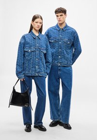 Traper jakne i traperice u plavoj nijansi s teksturiranim uzorkom. Model drži crnu kožnu torbu. Crne cipele upotpunjuju izgled.