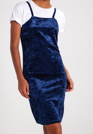 Tmavě modré hedvábné šaty na ramínka, přiléhavý střih a délka nad kolena; nošeny přes bílé tričko.