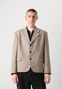 Blaser beige realizzato in tessuto testurizzato con colletto a bavero, tre bottoni neri e due tasche laterali. Indossato sopra un top nero con zip.