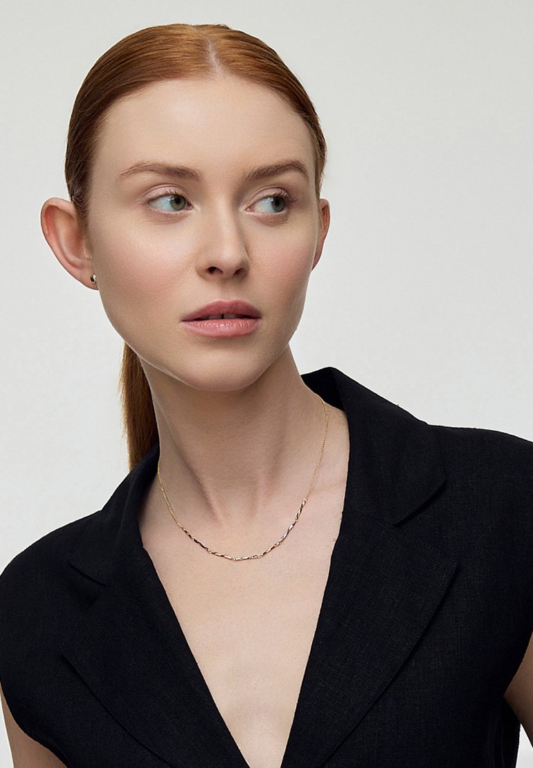 Złoty naszyjnik z łańcuszka o nieregularnych ogniwach, noszony na czarnym żakiecie. Modelka ma neutralny wyraz twarzy, włosy spięte z tyłu.
