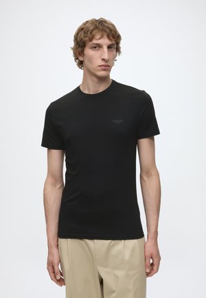 Jonge man met krullend haar die een strak zwart T-shirt en beige broek draagt, staand voor een effen witte achtergrond.