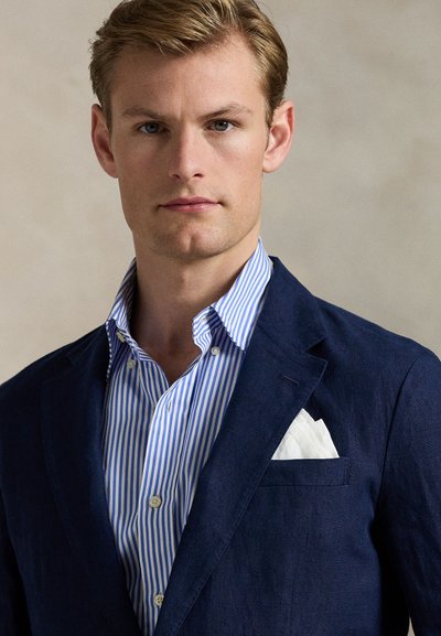 Navy blazer over a blue and white striped shirt; pocket square visible in the blazer pocket. Fabric appears crisp and smooth. Clean-cut design.
