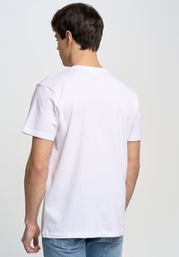 Camiseta de algodón blanca con cuello redondo y mangas cortas, vista desde la parte de atrás. Presenta un diseño simple sin patrones ni acentos visibles.