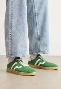 Tenisi din suede verde cu accente albe în dungi și șireți crem. Talpă din cauciuc. Asociați cu pantaloni din denim albastru deschis, cu manșetele ușor răsucite.