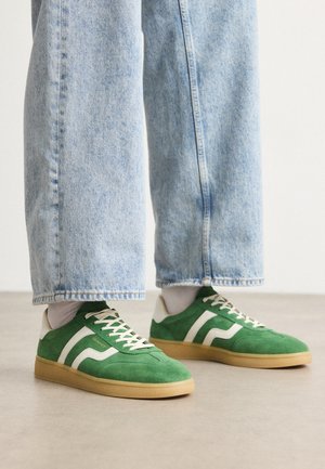 Tenisi din suede verde cu accente albe în dungi și șireți crem. Talpă din cauciuc. Asociați cu pantaloni din denim albastru deschis, cu manșetele ușor răsucite.