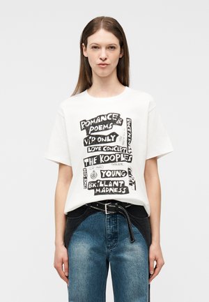 Γυναίκα με ίσια καστανά μαλλιά που φοράει λευκό t-shirt με μαύρο κείμενο και μπλε τζιν με μαύρες λεπτομέρειες στη ζώνη.