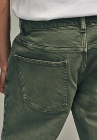 Khaki zöld farmernadrág klasszikus dizájnnal, dupla varrású derékpánttal, normál hátsó zsebbel és enyhe textúrával.