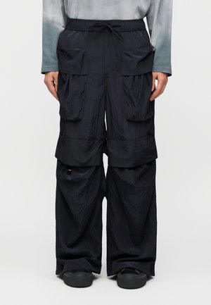 Χαλαρό μαύρο παντελόνι cargo με μεγάλες τσέπες με καπάκια και κορδόνι στη μέση, συνδυασμένο με μαύρα παπούτσια και γκρι μπλούζα μακρυμάνικη.