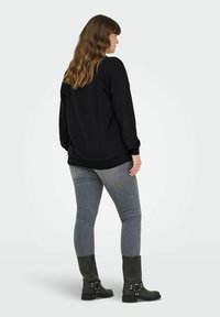 Sieviete ar viļņainiem brūniem matiem, uzvilkusi brīvu melnu džemperi, pelēkus šaurus džinsus un melnas zābaku līdz potītēm, stāv aiz muguras pret vienkāršu fona.