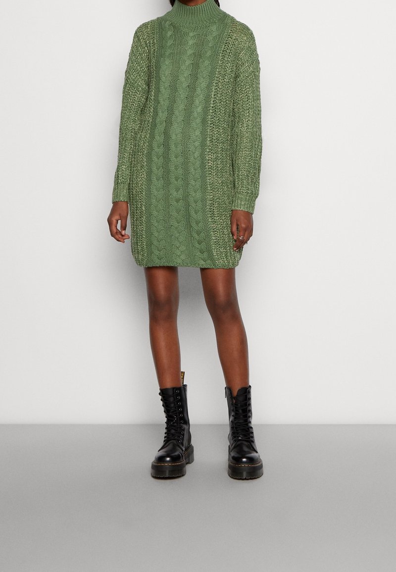 Personne portant une robe-pull à col roulé en maille torsadée verte et des bottes de combat noires à lacets, se tenant devant un fond clair uni.