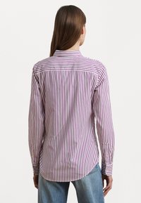 Camicia a maniche lunghe con righe bianche e viola, caratterizzata da un colletto classico, polsini con bottone e un orlo curvo. Il tessuto ha una consistenza liscia.
