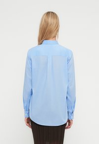 Svetlomodrá košeľa s dlhými rukávmi a golierom, s detailom záševku na chrbte, zaobleným lemom a manžetami na gombíky; spárovaná s tmavou plisovanou sukňou.