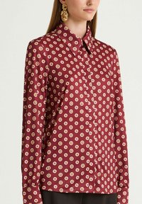 Burgundy button-up shirt featuring a geometric pattern in cream and pink. Long sleeves with button cuffs, smooth silk-like texture.
