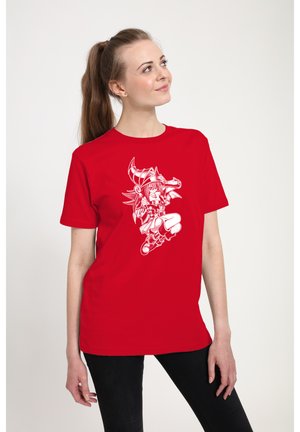 Ung kvinde med langt hår i hestehale, iført rød t-shirt med hvid detaljeret tegneseriefigur, der holder en lanse i en dynamisk positur.