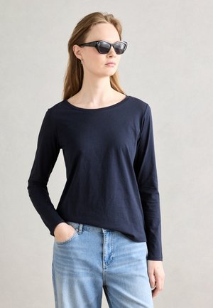 Marineblauwe longsleeve t-shirt van zachte katoen, ronde halslijn, gecombineerd met lichtblauwe spijkerbroek. Model draagt zwarte hoekige zonnebril.