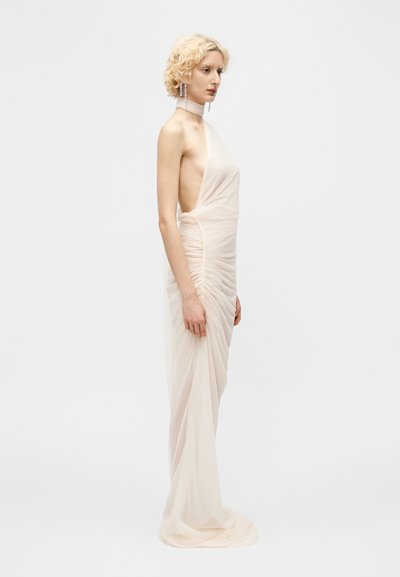 Woman with short blonde hair wearing a sleeveless, ruched, pale beige floor-length gown with a high neck and side cutouts, standing sideways.