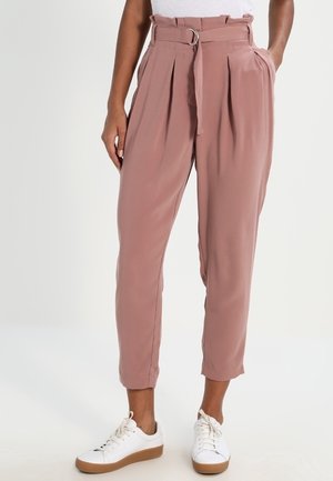 Pantalon plissé rose clair à taille haute, ceinture à nouer et poches latérales, associé à des baskets blanches et un haut ajusté. Texture lisse.