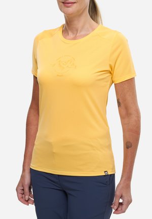 Femme portant un t-shirt jaune à manches courtes avec un logo embossé et un pantalon bleu marine, montrant des tatouages sur l'avant-bras et le poignet gauche.