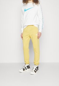 Vit sweatshirt med en stor blå Nike-logotyp, kombinerad med gula sweatpants med en liten vit Nike-logotyp, och svarta och vita sneakers.