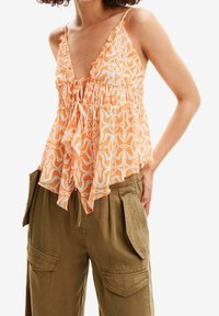 Haut sans manches à motif orange avec un détail de taille froncée, présentant un décolleté en V plongeant et un tissu léger et fluide, associé à un pantalon cargo olive.