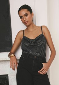 Haut noir sans manches avec un col drapé, fabriqué en tissu texturé et brillant ; associé à un pantalon taille haute noir et à un bracelet en or.