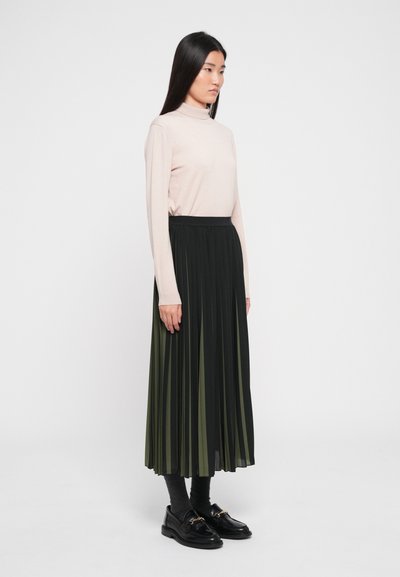 Pleated black and green skirt paired with a light pink long-sleeve top. Model stands sideways, wearing dark socks and black loafers.