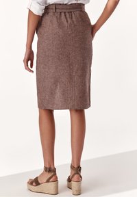 Nainen, jolla on päällään ruskea polvipitkä hame vyöllä, valkoinen ylös käärityt hihat paita ja ruskeat korkokengät, seisoo valkoisella lattialla.