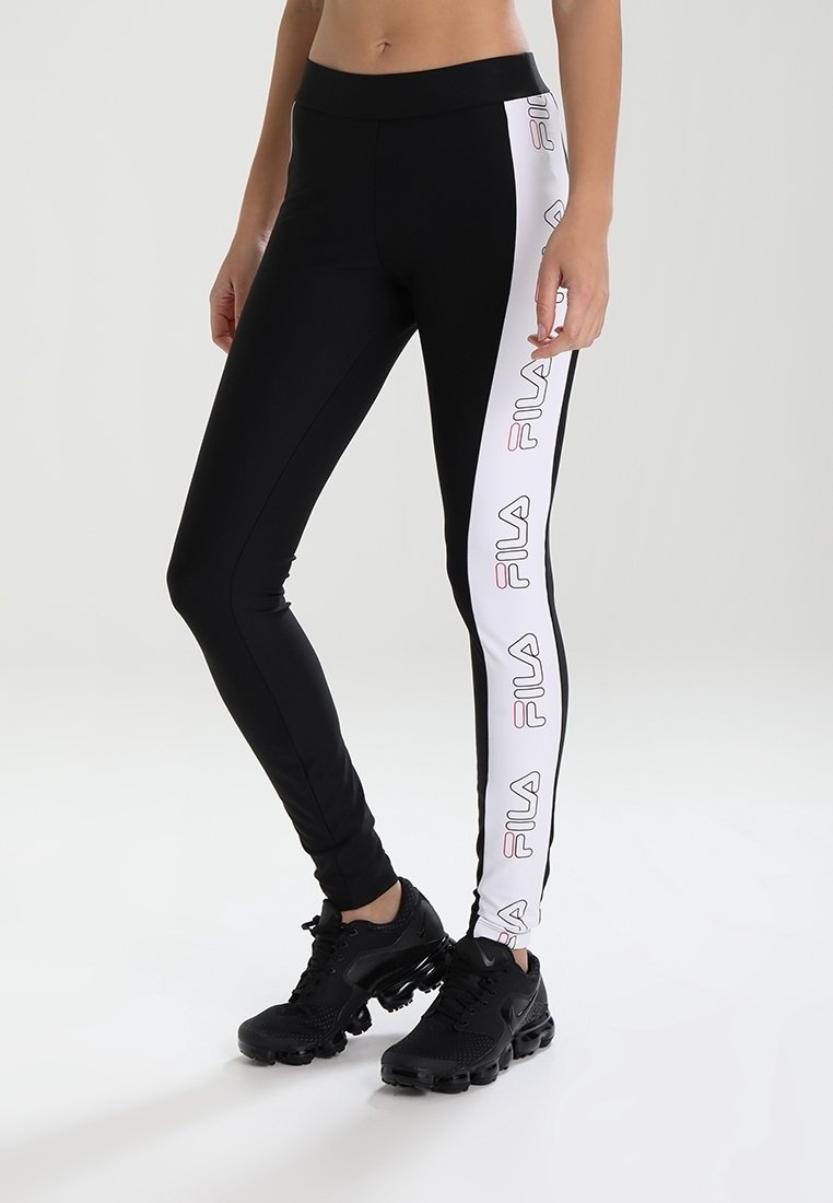 Kvinna som bär svarta leggings med vita sidopaneler som har upprepade FILA-logotyper, i kombination med svarta träningsskor.