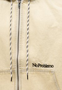Μπεζ φούτερ με φερμουάρ, μαύρα και λευκά κορδόνια με σχέδιο και μαύρο κεντητό κείμενο "No Problemo" πάνω από την μπροστινή τσέπη.