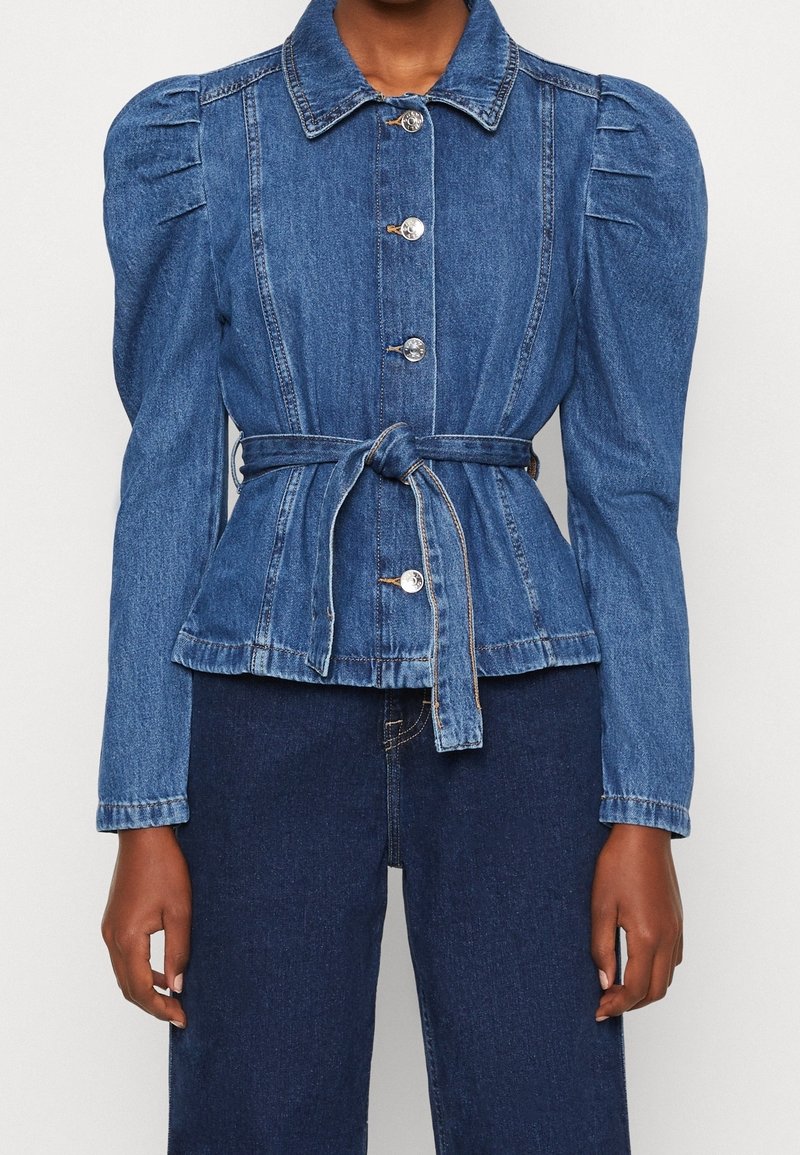 Džínová bunda s fitted siluetou, nabíranými rukávy, předním zapínáním na knoflíky a detaily vázacího pásku. Světle modrá barva s hladkou texturou.