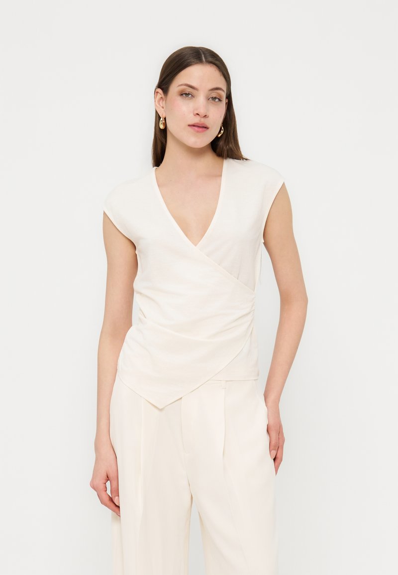 Νεαρή γυναίκα με μακριά καστανά μαλλιά, φοράει λευκό αμάνικο τοπ τύπου wrap και λευκό ψηλόμεσο παντελόνι που ταιριάζει, με ορατά χρυσά σκουλαρίκια.