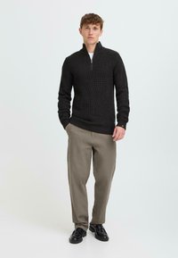 Donker geribd trui met een ritskraag, gecombineerd met lichtbeige broeken en zwarte schoenen. Eenvoudig ontwerp, textuurstof en neutrale kleuren.