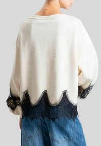 Felpa crema con bordo in pizzo nero all'orlo, caratterizzata da un design a forma di scallop. Vestibilità rilassata e maniche ampie. Indossata con jeans blu.