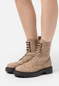 Beige mocka snörstövlar med brogue-detaljer, sidodragkedja och en tjock svart gummisula för förbättrat grepp.