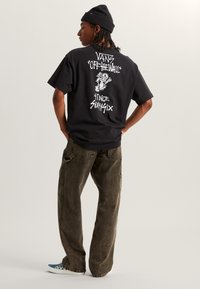 Camiseta de algodón negra con estampado gráfico blanco en la parte posterior, combinada con pantalones holgados beige y zapatillas a cuadros en azul y blanco.