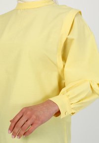 Blouse jaune à manches longues avec un col montant, des manches froncées et des poignets boutonnés. Fabriquée en tissu léger et lisse.