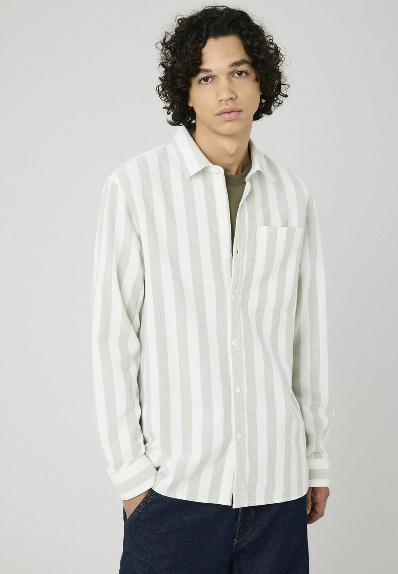 Jeune homme aux cheveux bouclés portant une chemise à rayures verticales blanches et gris clair sur une chemise verte, se tenant avec une main dans la poche.