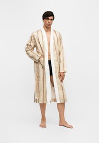 Homme debout pieds nus portant une robe de chambre beige rayée à manches longues, ouverte à l'avant avec des poches, par-dessus un short noir sur fond blanc.