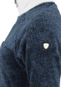 Marineblauwe fleece pullover met een gestructureerde, fluffy buitenkant, voorzien van een zilveren logo-patch op de mouw. Zacht, warm materiaal voor comfort.