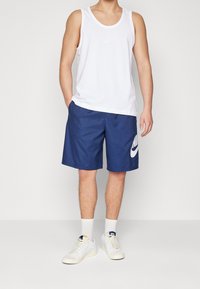 Biały bawełniany tank top z małym logo Nike, zestawiony z granatowymi szortami z dużym białym logo Nike. Białe trampki i skarpetki za kostkę.