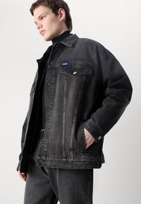 Black denim jacket with a relaxed fit, featuring button closure, two chest pockets, and a small blue logo patch on the left side.
