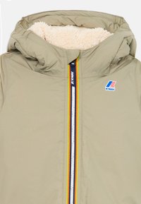 Veste à capuche beige avec doublure en polaire crème, fermeture éclair centrale avec bord multicolore, et logo K-Way bleu, rouge et blanc sur la poitrine.